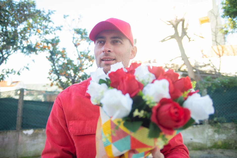 A male flower delivery courier dressed in a red jacket and matching red cap, standing outdoors with a backdrop of trees and a blue sky, holding a vibrant bouquet of fresh flowers wrapped in colorful p