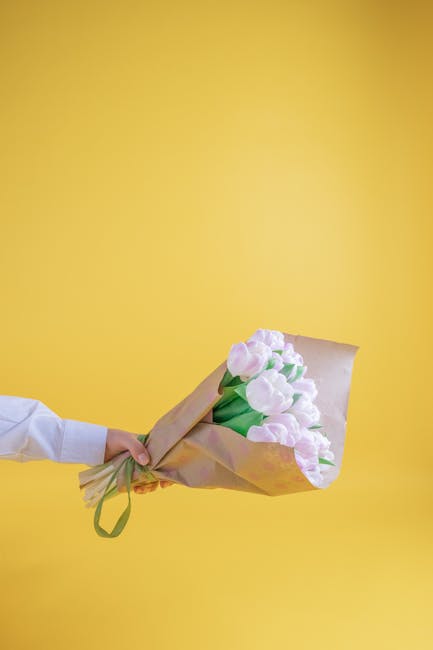 A person extending their arm to hold a bouquet of fresh, pastel pink tulips wrapped in brown kraft paper with a green ribbon, set against a vibrant yellow background. The tulips have smooth, delicate 
