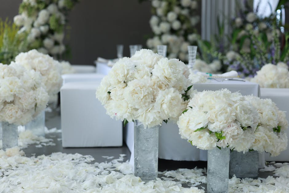 A floral arrangement featuring large, lush white hydrangea and peony blooms placed in tall, rectangular concrete vases as part of a wedding reception setup. The flowers are fresh, with textured petals