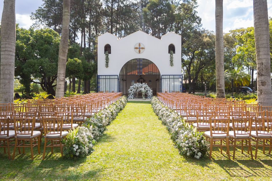 A view of an outdoor wedding setup in front of a white chapel with rounded arches and a cross symbol, surrounded by tall palm trees and lush greenery. The aisle is covered with bright green grass, fla
