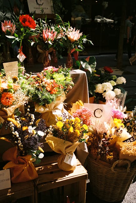 A display of various floral arrangements and bouquets on a wooden table and basket, featuring a mix of fresh flowers including pink lilies with long, slender petals, bright orange gerberas, yellow chr