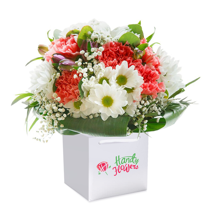 In the heart of Keston, a meticulously arranged floral bouquet takes centre stage, its lively palette and soft textures beautifully set off by a crisp, all-white square gift bag and a pristine backdrop. Leading the arrangement are abundant coral, peach, and lively red carnations-each one distinguished by deeply ruffled petals that create a sense of light movement and fullness. Interspersed between the larger blooms, pristine white daisy-like chrysanthemums with vibrant lime-green centers lend a refreshing brightness, reminiscent of Keston's tranquil parklands and nature walks. Sprigs of baby's breath are woven elegantly throughout, imparting a gentle, cloud-soft effect that inspires feelings of lightness and calm. Peering from the left, subtle purple-tipped buds add intrigue and evoke the promise of flowers yet to blossom-much like the evolving spring meadows just beyond Fox Hill. A collar of broad, glossy leaves in rich green shades provides essential structure, echoing the local landscape's lush character. Practical white ribbon handles make the minimalist gift bag easy to carry or present, perfectly suited for celebrations across Keston, whether welcoming a new neighbor or marking a personal milestone. Every detail reflects artistry and fresh, heartfelt intent-a bouquet crafted by skilled local hands for moments of connection and joy.