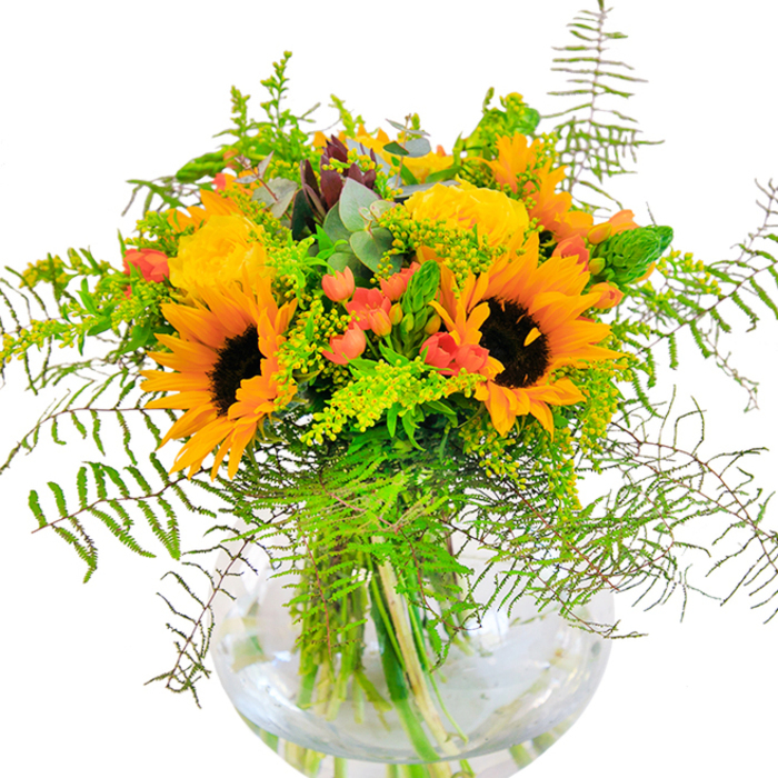 First impressions of this bouquet bring to mind the lively energy of Keston's summer gardens: golden sunflowers stand tall and wide, petals spreading in generous layers around rich brown centres, exuding a sense of optimism that feels at home near the historic ponds of Keston village. Nestled close are plush, yellow garden roses, their tightly layered blooms reminiscent of carefully tended local rose beds, adding a note of classic charm to the display. Sparkling between the larger flowers, ruby-red Hypericum berries glisten with vitality, their glossy skins set against the soft, matte finish of petals and lush, deep green foliage. Fern fronds and airy filler cascade outwards, their movement and texture evoking moments spent wandering along the trails of Hayes Common or relaxing on the edges of Keston's woodlands. Goldenrod and slender spikes provide playful height and intricate detail, while the aroma of eucalyptus mingles gently-a subtle, refreshing finish. The entire bouquet is thoughtfully arranged in a fishbowl-style, clear glass vase, its water magnifying the beauty of every stem and leaf. This composition is a heartfelt nod to the artistry and natural abundance found in Keston, perfect for birthdays, anniversaries, or any joyful occasion that calls for flowers with depth, character, and a sense of local pride. Arrangements like this, prepared by Florist Keston, celebrate the stories and traditions cherished by the community.