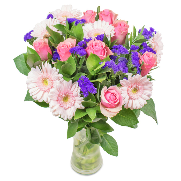 A radiant, meticulously arranged bouquet rests within a slender clear glass vase atop a crisp white surface. The focal point is a collection of 7-9 soft and mid-pink roses, displaying every stage from tightly furled buds to partially open blooms, their gentle curves forming inviting spirals. They are surrounded by about 5-7 pale pink Gerbera daisies, distinguished by their large, rounded faces and fine, radiating petals encircling creamy near-white centres. Artfully distributed among the blooms are vibrant purple clusters of statice, adding texture and a vivid pop that captures the eclectic charm of Keston's countryside gardens. Lush, deep green foliage forms a generous base, recalling the abundant greenery of Keston village and its surrounding commons. The clear water and crossed stems within the glass suggest both refreshing simplicity and freshly picked quality-a hallmark of Florist Keston's commitment to mindful floristry. Ideal for birthdays, anniversaries, Mother's Day, or as a beautiful accent in a local home or office, this arrangement conveys celebration, grace, and a touch of Keston's serene rural beauty.